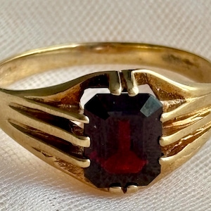 May include: A gold ring featuring a central emerald-cut dark red gemstone. The band has a polished finish and includes three distinct grooved lines on each side that lead up to the prongs securing the rectangular stone in a prong setting.