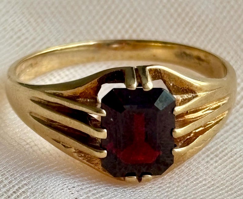 May include: A gold ring featuring a central emerald-cut dark red gemstone. The band has a polished finish and includes three distinct grooved lines on each side that lead up to the prongs securing the rectangular stone in a prong setting.