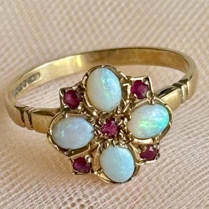 May include: A gold ring featuring a floral cluster design. The centre of the ring is set with four oval-shaped white opals arranged in a cross pattern, interspersed with four small round red rubies. The band is made of polished yellow gold.