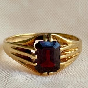 May include: A gold ring featuring a rectangular, emerald-cut dark red gemstone set in the center. The band is crafted from polished yellow gold and includes three distinct, parallel metal prongs on each side that secure the stone in place.