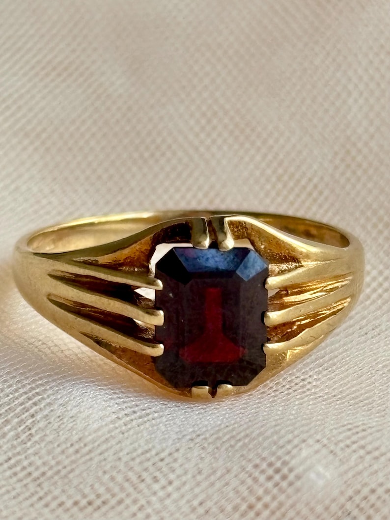 May include: A gold ring featuring a rectangular, emerald-cut dark red gemstone set in the center. The band is crafted from polished yellow gold and includes three distinct, parallel metal prongs on each side that secure the stone in place.