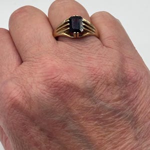 May include: A gold ring featuring a rectangular, deep red gemstone set in a prong setting. The band of the ring is constructed with three distinct, parallel gold bars on each side that converge as they meet the central stone setting.