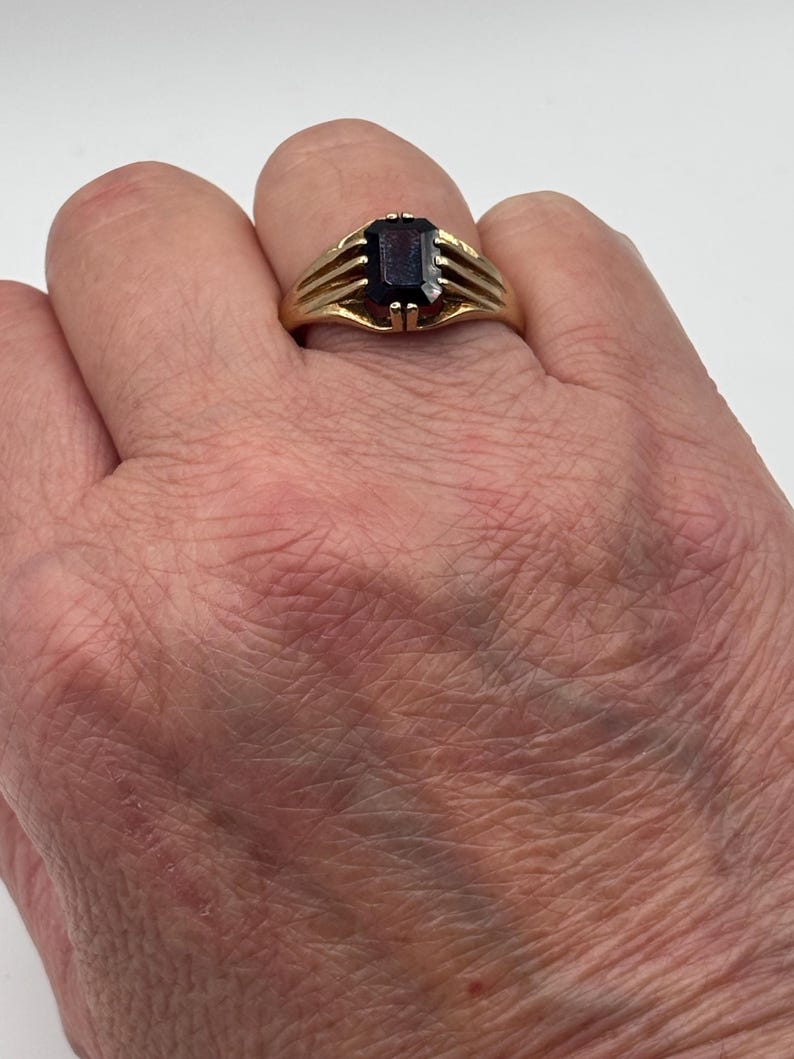May include: A gold ring featuring a rectangular, deep red gemstone set in a prong setting. The band of the ring is constructed with three distinct, parallel gold bars on each side that converge as they meet the central stone setting.