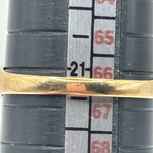 May include: A gold ring positioned on a black ring mandrel. The ring rests directly over the number 21 on the sizing scale, which is printed in black on the gray surface of the mandrel. The metal band appears smooth and polished in appearance.