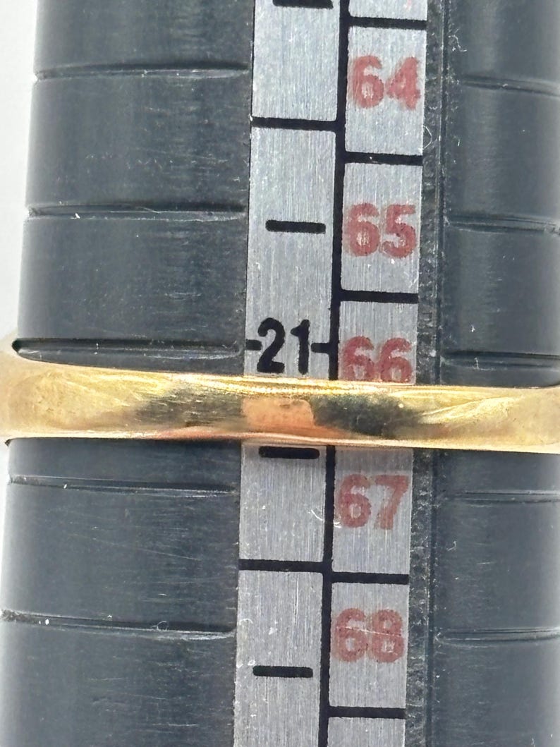 May include: A gold ring positioned on a black ring mandrel. The ring rests directly over the number 21 on the sizing scale, which is printed in black on the gray surface of the mandrel. The metal band appears smooth and polished in appearance.