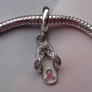Könnte beinhalten: Ein silbernes Armband mit einem silbernen Anhänger in Form eines weißen Flip-Flops mit einem rosa Band. Der Flip-Flop ist mit klaren Strasssteinen verziert.