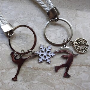 May include: White braided keychain with two silver key rings. Each key ring has a silver charm of a figure skater and a snowflake. One snowflake charm is white with a silver outline, and the other is silver with a snowflake design.