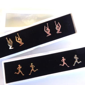 Puede incluir: Dos pares de pendientes de botón en cajas blancas con inserciones de terciopelo negro. Un par presenta siluetas de gimnastas, el otro corredores. Los pendientes son dorados y plateados, mostrando figuras atléticas en movimiento.