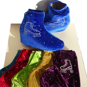 May include: Royal blue velvet roller skate boot covers with rhinestone embellishments. The covers feature a rhinestone design of a roller skate. Additional covers in red, green, gold, and purple are also visible.