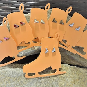May include: Assortment of ice skate charm earrings displayed on orange cardboard cutouts shaped like ice skates. The earrings are silver with different coloured enamel accents.