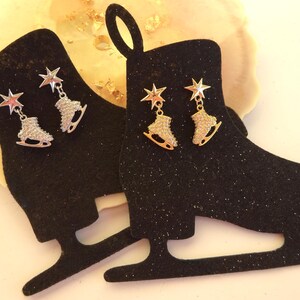 May include: Black glitter ice skate-shaped display stands with star and ice skate earrings. One pair of earrings is silver with red star accents, the other is gold. The earrings are dangling from the top of the skate.