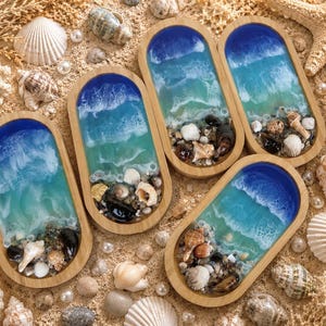 Könnte beinhalten: Sechs Holztabletts mit einem Harz-Ozean-Design, das blaue und türkisfarbene Wellen, Muscheln und Sand zeigt. Die ovalen Tabletts sind von verschiedenen Muscheln, Seesternen und Perlen umgeben, was ein Küstenmotiv erzeugt.