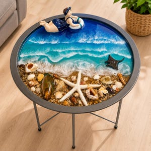 Pode incluir: Uma mesa lateral redonda com uma cena oceânica de resina com ondas brancas, uma área de praia arenosa cheia de várias conchas, uma grande estrela-do-mar branca, uma figura de raia preta e uma pequena estatueta de uma pessoa com boia.