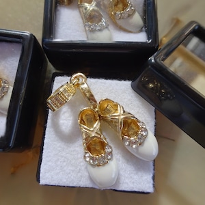 Pode incluir: Um par de pingentes de sapato de bailarina em tom dourado com esmalte branco e strass transparente. Os pingentes são exibidos em uma pequena caixa preta.
