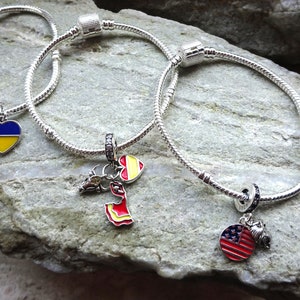 Puede incluir: Tres pulseras de plata con dijes de banderas de diferentes países. La primera pulsera tiene un dije de corazón azul y amarillo, la segunda tiene un dije de corazón rojo y amarillo, y la tercera tiene un dije circular rojo, blanco y azul con un pequeño dije de águila.