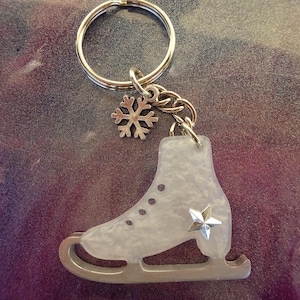 May include: A silver-coloured keyring with a translucent ice skate charm, a star accent, and a snowflake charm. The keyring has a silver ring and chain. The background is a blend of purple, grey, and white.
