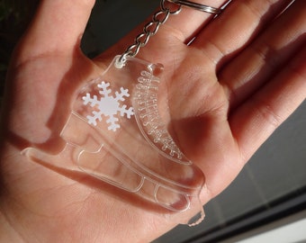 May include: A clear plastic ice skate keychain with a white snowflake design. The keychain is attached to a silver chain.