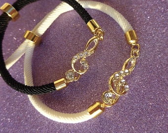 Peut inclure: Deux bracelets, un noir et un blanc, avec des accents dorés. Chaque bracelet est orné d'un charm en forme de clé de sol doré orné de cristaux transparents. Les bracelets sont posés sur un fond violet scintillant.