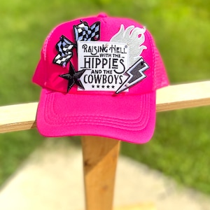 May include: A pink trucker hat with a black and white checkered patch, a white flame patch, a black star patch, and a white patch with the text "Raising Hell with the Hippies and the Cowboys" in black.