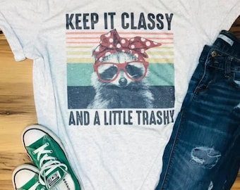 May include: A gray t-shirt with a retro-style graphic of a raccoon wearing sunglasses and a red bandana with white polka dots. The text "Keep it Classy and a Little Trashy" is printed on the shirt.