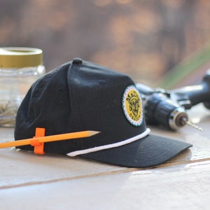 May include: A black baseball cap with a white brim and a yellow and green logo that says "Made Design". A pencil is clipped to the cap with an orange clip.