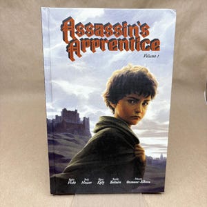 May include: A hardback book titled "Assassin's Apprentice, Volume 1" shows a young boy in a hooded cloak. The cover art includes a castle and a cloudy sky. The title is in orange and brown lettering.
