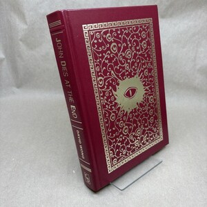 Puede incluir: Un libro de tapa dura burdeos titulado "John Dies at the End" de David Wong. La portada presenta detalles dorados, incluido un diseño en forma de ojo. El lomo muestra el título y el nombre del autor.