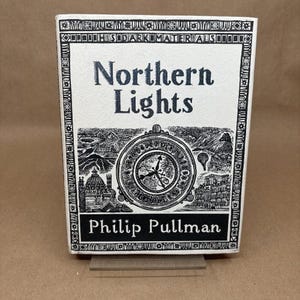 Peut inclure: Un livre à couverture rigide intitulé "Northern Lights" de Philip Pullman. La couverture présente une illustration en noir et blanc d'une boussole, de montagnes et de bâtiments. Le titre est en gras.