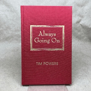May include: A hardcover book with a red cover and gold lettering. The title "Always Going On" is displayed in a gold rectangular frame. The author's name, "TIM POWERS," is below the title. The book has a textured appearance.