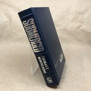 Könnte beinhalten: Ein marineblaues Hardcover-Buch mit dem Titel "SUBMERGED" in silberner Schrift. Der Name des Autors, Thomas F. Monteleone, ist auf dem Buchrücken aufgedruckt. Das Buch wird auf einem transparenten Acrylständer präsentiert.