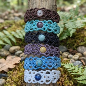 May include: A stack of six macrame bracelets featuring intricate knotted patterns and a single central gemstone in each. The bracelets are colored brown, turquoise, black, purple, blue, and white, each with a unique stone at the center.