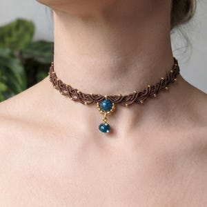 May include: A brown macrame choker necklace featuring a woven cord design accented with small gold-toned beads. The centre of the choker displays a round blue stone set in gold, with a matching blue and white striped bead dangling from it.
