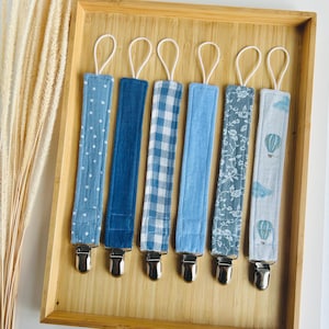 Può includere: Una collezione di sei clip per ciuccio per bambini in vari modelli blu, tra cui pois, denim tinta unita, quadretti e motivi floreali. Ogni clip ha un fermaglio in metallo e un anello bianco, esposti in un vassoio di legno.