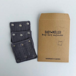 Puede incluir: Tres extensores de body de tela gris con cierres a presión apilados junto a un sobre de papel marrón. El sobre tiene el texto "BABY MÖLLER Body Suit extender" y un dibujo de un body de bebé. Las palabras "HANDMADE IN SWEDEN" están impresas en la parte inferior.