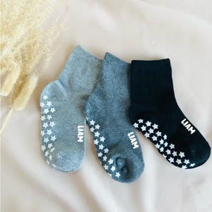 Könnte beinhalten: Drei Paar graue und schwarze Kniestrümpfe mit weißen Sternengriffen an der Unterseite. Die Socken haben den Namen "LIAM" aufgedruckt.