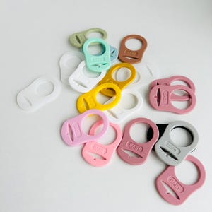 Puede incluir: Una colección de clips para chupetes de bebé en varios tonos, incluyendo rosa, amarillo, verde y blanco. Cada clip tiene un diseño circular con un lazo y una sección rectangular con la palabra "BABY" impresa.