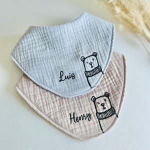 May include: Two baby bibs, one light blue and one beige, with a bear illustration and the names "Luis" and "Henry" in black script. The bibs are made of a textured fabric.