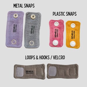 May include: Rectangular fabric straps labeled BABY M&Ouml;LLER HANDMADE IN SWEDEN. Top left: two purple and gray straps with metal snaps. Top right: two pink and yellow straps with plastic snaps. Bottom: two brown straps with hook and loop fasteners.