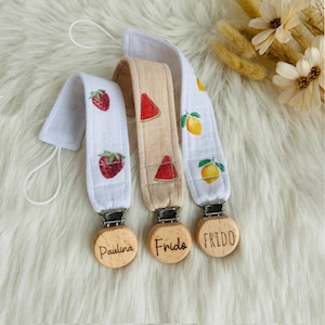 Puede incluir: Tres clips para chupete de bebé con etiquetas de nombre de madera. Uno tiene fresas, otro rodajas de sandía y el tercero limones. Los nombres "Paulina", "Frida" y "Frido" están grabados en las etiquetas de madera.