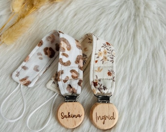 Puede incluir: Dos clips de tela para chupete con bucles elásticos blancos y discos de madera redondos. Un clip presenta un estampado de leopardo marrón y el otro un patrón floral. Los discos de madera tienen grabados los nombres Sakina e Ingrid.