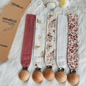 May include: Five baby pacifier clips with wooden beads and fabric straps in various patterns and colors, including burgundy, floral, and white. The pacifier clips are displayed on a fluffy white surface, with a cardboard package that reads "BABY-MÖLLER" in the background.