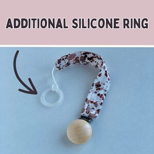 Könnte beinhalten: Ein Schnullerhalter für Babys mit einer Holzperle und einem Stoffband mit braunen Flecken auf weißem Grund. Das Bild zeigt einen transparenten Silikonring und den Text "ADDITIONAL SILICONE RING".