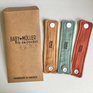 Pode incluir: Conjunto de três extensores de babador de tecido castanho, verde e vermelho com botões brancos. Cada extensor tem o texto "BABY MÖLLER" impresso nele. Os extensores são embalados num envelope de papel castanho com o texto "BABY MÖLLER Bib extender" e "HANDMADE IN SWEDEN" impresso nele.