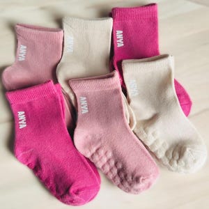 Könnte beinhalten: Sechs Paar Knöchelsocken in Rosa- und Cremetönen. Jede Socke trägt das Wort "ANYA" in Weiß. Die Socken haben strukturierte Sohlen für besseren Halt.