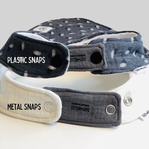 May include: Two baby bibs stacked vertically. The top bib is dark gray with white spots and black plastic snaps. The bottom bib is white and dark gray with silver metal snaps. Both bibs feature a small white tag labeled BABY-M&Ouml;LLER.