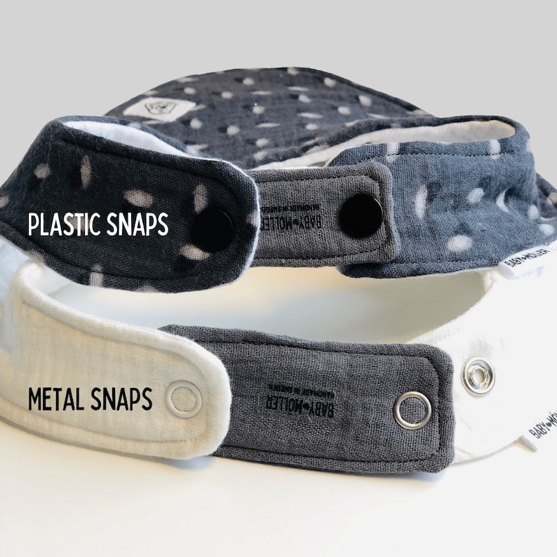 May include: Two baby bibs stacked vertically. The top bib is dark gray with white spots and black plastic snaps. The bottom bib is white and dark gray with silver metal snaps. Both bibs feature a small white tag labeled BABY-M&Ouml;LLER.