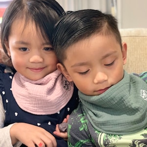 Puede incluir: Dos ni&ntilde;os con baberos. Uno lleva un babero rosa y un vestido azul marino con lunares blancos. El otro lleva un babero verde y una camisa verde y gris con un estampado de dinosaurio.