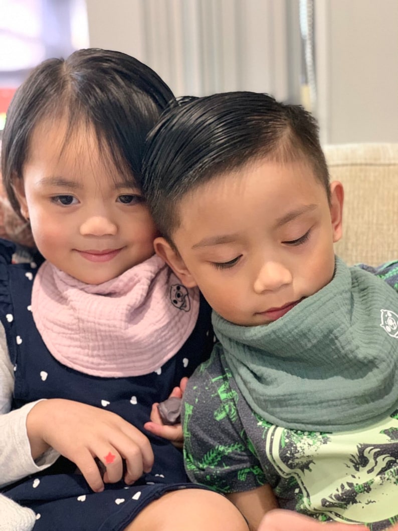 Puede incluir: Dos ni&ntilde;os con baberos. Uno lleva un babero rosa y un vestido azul marino con lunares blancos. El otro lleva un babero verde y una camisa verde y gris con un estampado de dinosaurio.