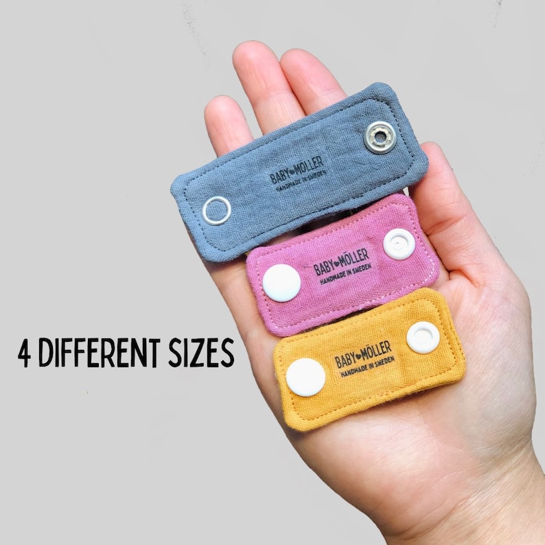 May include: Three rectangular fabric strap extenders in blue, pink, and yellow, each featuring two white plastic snap buttons and the black printed text BABY M&Ouml;LLER HANDMADE IN SWEDEN. The text 4 DIFFERENT SIZES is visible on the left side.