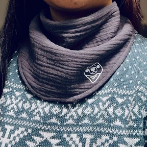 Puede incluir: Un pa&ntilde;uelo gris texturizado tipo babero con un logotipo de cara de oso de dibujos animados blanco. El pa&ntilde;uelo se lleva sobre un su&eacute;ter azul y blanco estampado. El pa&ntilde;uelo est&aacute; hecho de una tela suave y transpirable.